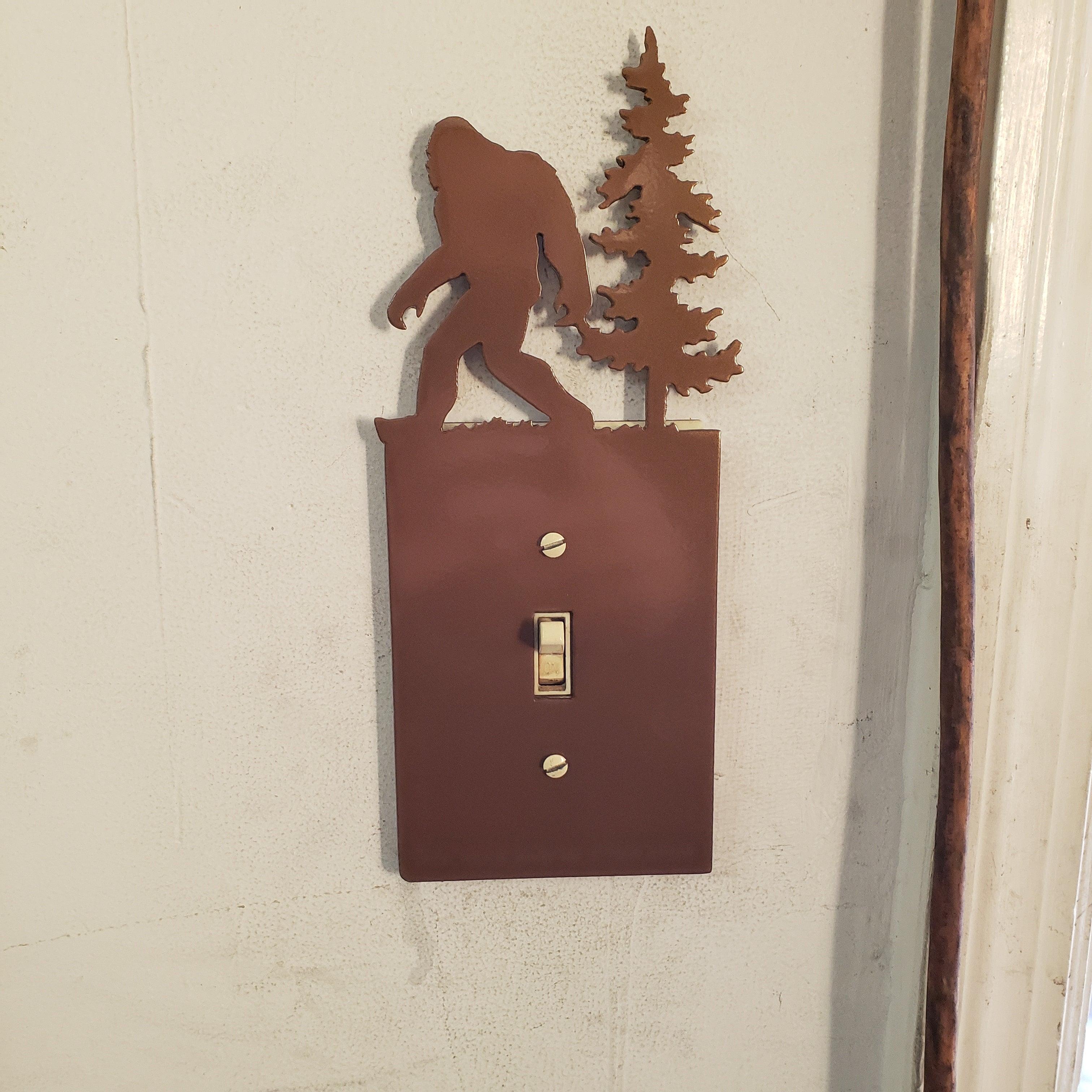Metallic bigfoot design on a light switch cover with trees against a beige wall.