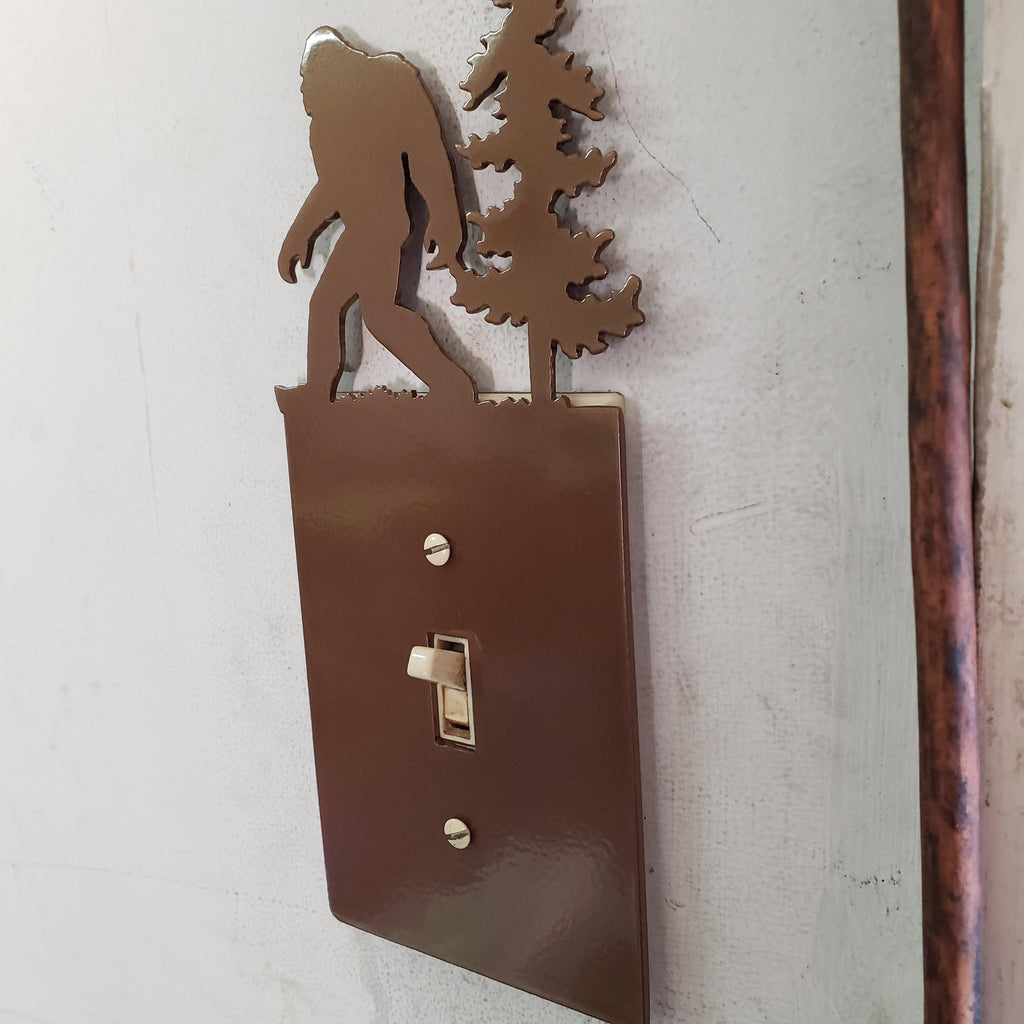 Decorative metal light switch cover with a silhouette of bigfoot and trees on a white wall.