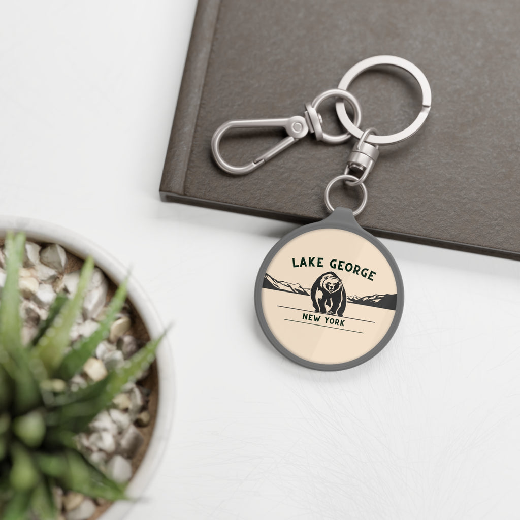 Round keychain with 'Lake George, New York' design on a white surface with a plant and book in the background.
