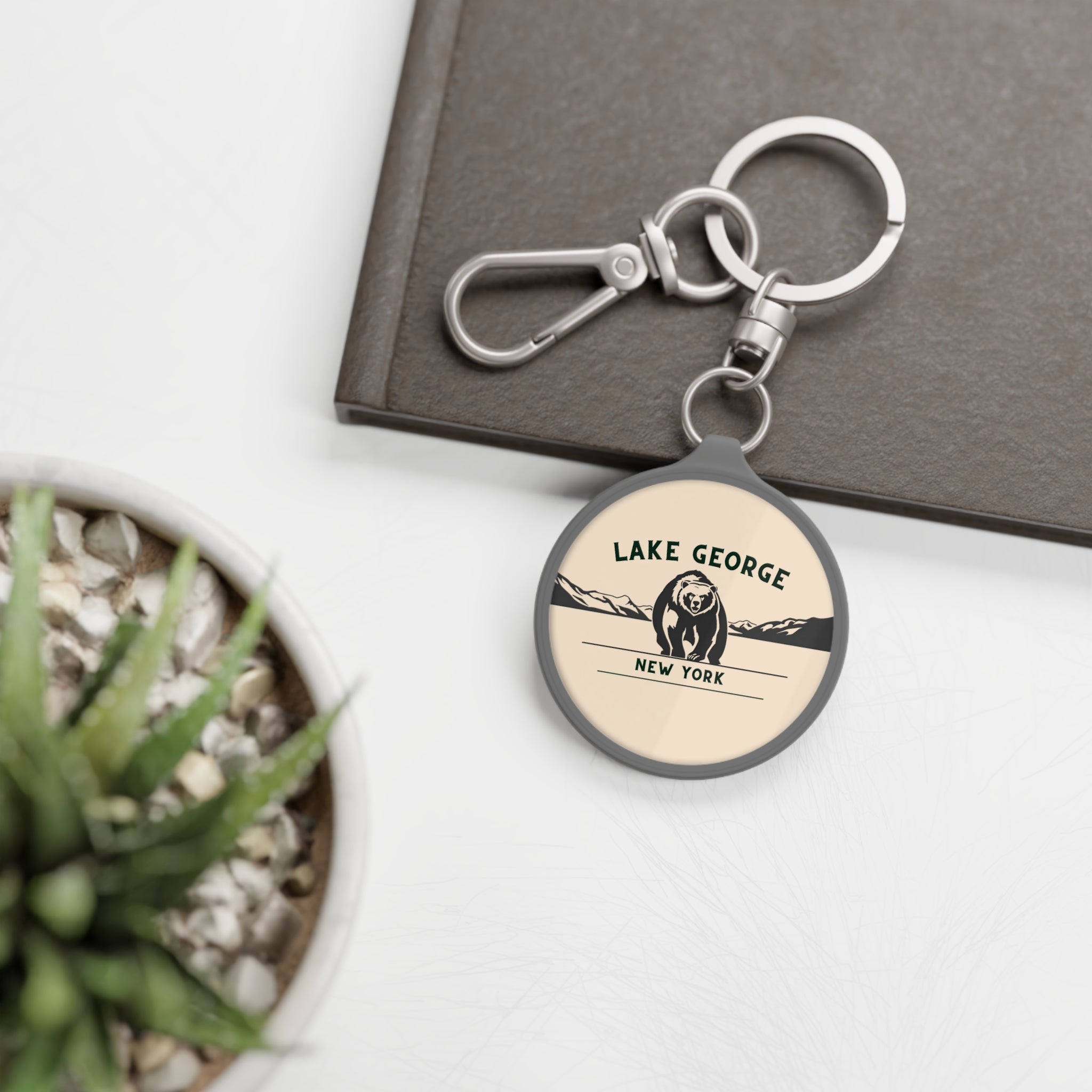 Round keychain with 'Lake George, New York' design on a white surface with a plant and book in the background.