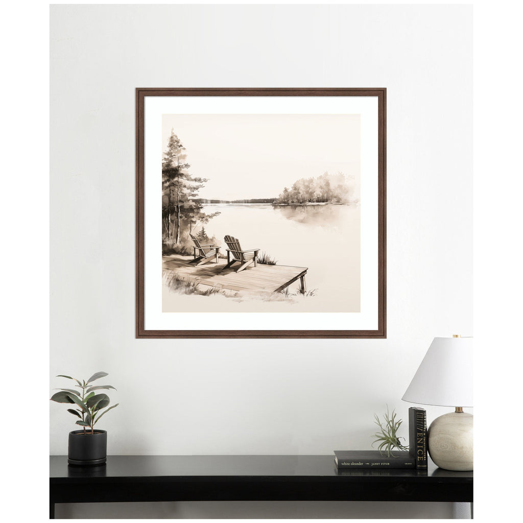 Framed artwork of a lakeside scene with Adirondack chairs on a white wall above a desk.
