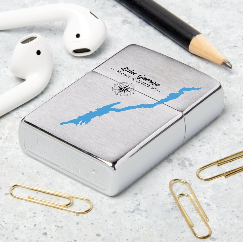 A silver chrome Zippo-style lighter with a brushed metal finish, featuring a blue map graphic and the printed name 'Luke George' with an address and compass rose. The lighter is styled on a light gray speckled surface with white earbuds, a black pencil, and gold paperclips.